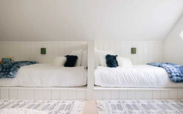 Built-in twin beds with white paneling, soft neutral bedding, and sloped ceiling creating a cozy shared kids sleeping area